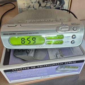 Può includere: Una radiosveglia Sony Dream Machine argentata con un display digitale verde che mostra le 8:59. La radio ha pulsanti per l'impostazione dell'ora, la sveglia e la radio. La scatola sottostante riporta la scritta "Triple visualización de horarios".