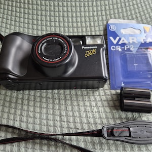 Peut inclure: Un appareil photo Panasonic Zoom noir avec un objectif 38-80mm, une sangle de cou noire avec des coutures rouges, une pile Varta CR-P2 dans son emballage bleu et blanc d'origine, et un composant de batterie cylindrique noir à côté du boîtier.