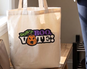 May include: A natural-colored canvas tote bag featuring a graphic print that reads "DON'T BOO. VOTE!" with an orange jack-o'-lantern illustration replacing the letter "O" in the word "BOO" and the "O" in the word "VOTE".