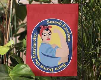 May include: A red rectangular garden flag featuring a central illustration of a person with a red polka dot headscarf and blue shirt flexing their arm. The text "Smash Fascism. Burn Down the Patriarchy." is printed in yellow within a blue oval.