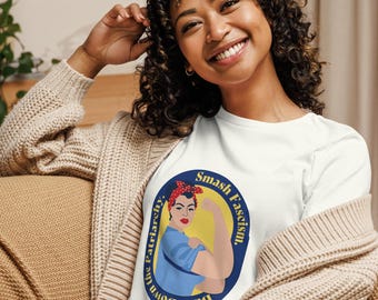 May include: A white t-shirt featuring a circular graphic of a person with a red polka dot headband flexing their arm. The text surrounding the graphic reads, "Smash Fascism, Burn Down the Patriarchy." The shirt is worn under a beige knit cardigan.
