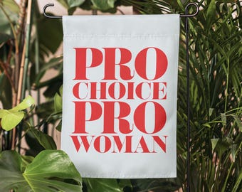 May include: A rectangular white garden flag featuring the red text PRO CHOICE PRO WOMAN in a bold serif font. The flag is suspended from a black metal stand, displaying the message clearly across the center of the fabric surface.