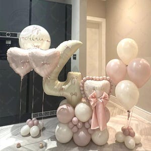 Pode incluir: Um conjunto de balões rosa e brancos, incluindo balões em forma de coração e número, para uma celebração. Os balões são decorados com laços e o nome "Alria".