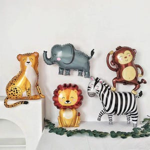 May include: A collection of animal-shaped foil balloons, including a cheetah, elephant, monkey, zebra, and lion. The cheetah is gold with black spots, the elephant is gray with pink cheeks, and the monkey is brown. The zebra has black and white stripes.