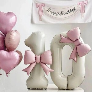 Könnte beinhalten: Eine Ansammlung von rosa herzförmigen und runden Luftballons, zusammen mit großen, cremefarbenen Zahlenballons in Form einer "1" und einer "0", die jeweils mit einer rosa Schleife verziert sind. Im Hintergrund ist ein Banner mit der Aufschrift "Happy Birthday" zu sehen.