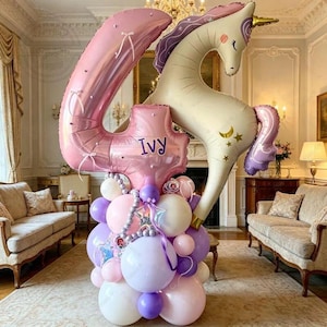 Könnte beinhalten: Eine Partydekoration mit einem großen, rosafarbenen Ballon in Form der Zahl Vier und einem Einhorn-Ballon. Der Ballon mit der Zahl Vier hat weiße Schleifen und den Namen "Ivy" auf einem kleineren Ballon. Das Einhorn ist cremefarben mit lila Mähne und Schweif.