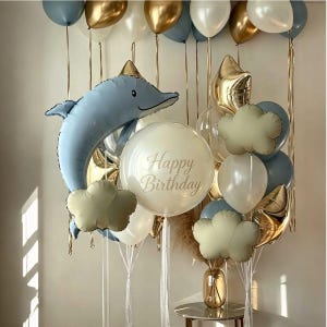 Könnte beinhalten: Ein Ballonarrangement mit einem großen blauen delfinförmigen Folienballon, einem zentralen transparenten Ballon mit dem goldenen Schriftzug Happy Birthday sowie verschiedenen runden und wolkenförmigen Ballons in Blau, Weiß, Gold und Salbeigrün.