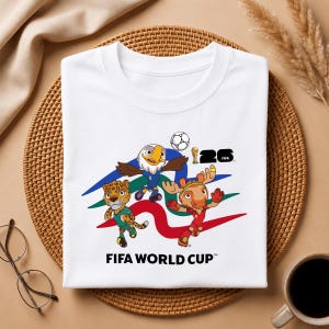 May include: White t-shirt featuring a colorful graphic of three cartoon mascots, a soccer ball, and the text "FIFA WORLD CUP". The design includes an eagle, a jaguar, and a deer-like character. The shirt also has the number "26" and a trophy.