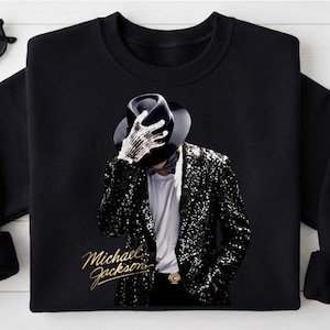 May include: A black crewneck sweatshirt featuring a graphic print of a figure wearing a black fedora, a white t-shirt, and a black sequined blazer. The figure wears a sparkling glove and holds the hat. The text Michael Jackson is in gold.