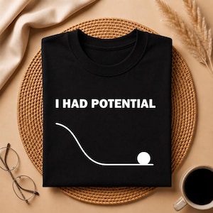 Può includere: T-shirt nero girocollo con la scritta bianca "I HAD POTENTIAL" sopra una grafica a linea bianca che raffigura una palla che rotola giù per un pendio. La maglietta è esposta su un tappetino intrecciato.