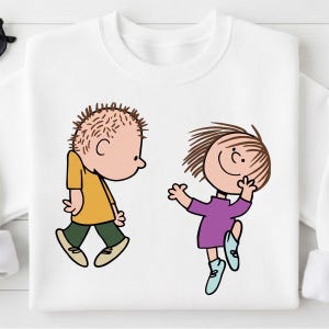 May include: White sweatshirt featuring cartoon illustrations of two children. One child is wearing a yellow shirt and green pants, the other is wearing a purple dress and blue shoes. The characters are in a playful pose.