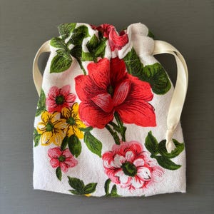 Könnte beinhalten: Ein kleiner beigefarbener Stoffbeutel mit Kordelzug und einem lebendigen Blumenmuster aus großen roten, gelben und rosa Blüten sowie grünen Blättern. Der Beutel ist oben mit einem cremefarbenen Satinband an beiden Seiten verschlossen.