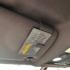 Könnte beinhalten: Ein schwarzer Sonnenblendenclip für das Auto, der eine Karte mit dem Text "TREVENA" hält. Die Karte hat ein gelbes und grünes Design. Die Sonnenblende ist grau und aus einem strukturierten Stoff. Der Innenraum eines Autos ist sichtbar.