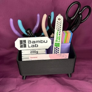 May include: A black, rectangular desk organizer with a variety of tools, including scissors, pliers, and a white tool with the text "Bambu Lab." The organizer also holds small, colorful rectangular blocks and business cards.