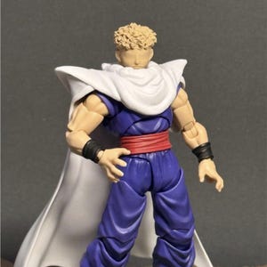 Puede incluir: Figura de acción detallada de un personaje con un atuendo morado y azul, con una capa blanca y un cinturón rojo. La figura tiene piel bronceada, cabello rubio y muñequeras negras. La figura está de pie.