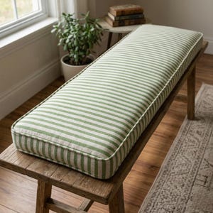 May include: A rectangular bench cushion with a green and white striped pattern. The cushion is placed on a rustic wooden bench. The cushion has a green border. The bench is in a room with a window, a plant, and a small side table.