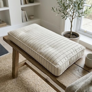 May include: A rectangular cushion with a cream-colored fabric and thin, dark gray vertical stripes. The cushion is placed on a rustic wooden bench. The cushion has a white border.