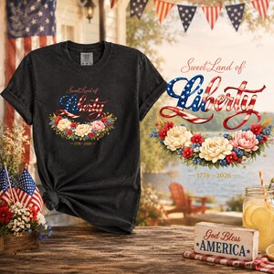 May include: Dark gray t-shirt with the text "Sweet Land of Liberty" in red, white, and blue, with a floral wreath design. The shirt also has the date "1776-2026" printed below the floral wreath.