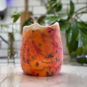 Puede incluir: Un jarrón naranja afieltrado hecho a mano con detalles en rosa y negro. El jarrón tiene una superficie texturizada y una forma orgánica única. El borde superior es de un color blanquecino más claro. Este artículo decorativo es adecuado para la decoración del hogar.
