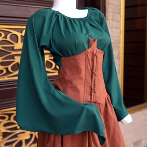 Op de afbeelding: Een groene top met lange, uitlopende mouwen en een vierkante halslijn, gecombineerd met een bruin korset en rok. Het korset heeft bruine veters. De outfit doet denken aan historische kleding.