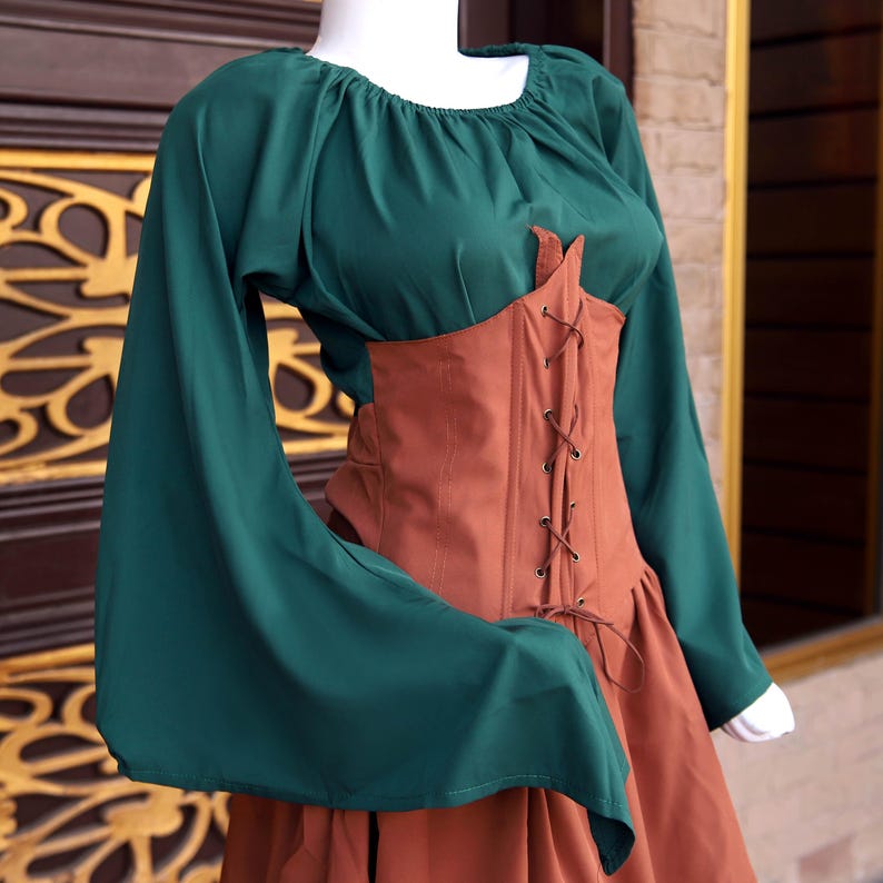 Op de afbeelding: Een groene top met lange, uitlopende mouwen en een vierkante halslijn, gecombineerd met een bruin korset en rok. Het korset heeft bruine veters. De outfit doet denken aan historische kleding.