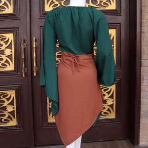 Op de afbeelding: Een tweedelige outfit met een donkergroene top met lange mouwen en een roestkleurige wikkelrok. De top heeft een ronde halslijn en uitlopende mouwen. De rok wikkelt zich om de taille en wordt aan de voorkant vastgebonden, waardoor een gelaagd effect ontstaat.