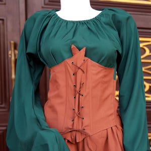Op de afbeelding: Een groene boeren top met lange mouwen en een roestkleurig korset. Het korset heeft een veter aan de voorkant en is bevestigd aan een bijpassende roestkleurige rok. De outfit wordt op een mannequin getoond.