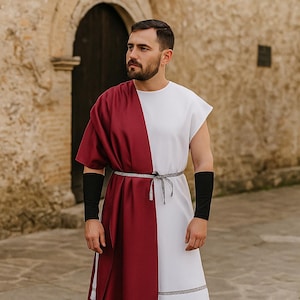 Puede incluir: Una túnica de estilo romano, dividida verticalmente con tela blanca y burdeos. La prenda se ciñe a la cintura con un cinturón de cuerda plateada. Se usan mangas negras en los brazos. El fondo es un edificio de piedra con una puerta arqueada.