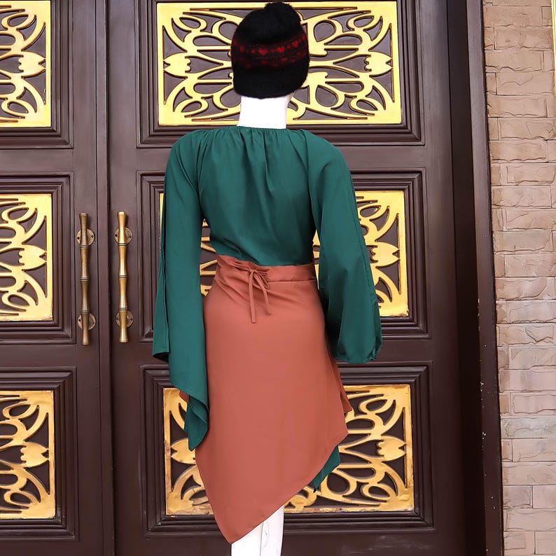 Op de afbeelding: Een groene top met lange, uitlopende mouwen en een bruine wikkelrok. De rok heeft een striksluiting in de taille. Een zwarte gebreide muts met rode accenten staat op het hoofd van de mannequin. De outfit wordt gepresenteerd voor een bruine deur.
