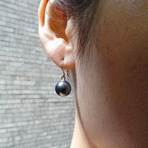 May include: A close-up of a person's ear with a single black pearl earring. The earring is a simple design with a small gold hook.
