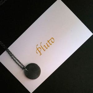 Puede incluir: Un collar de cadena de plata con un colgante redondo negro que dice "Pluto" en escritura dorada en una tarjeta blanca.