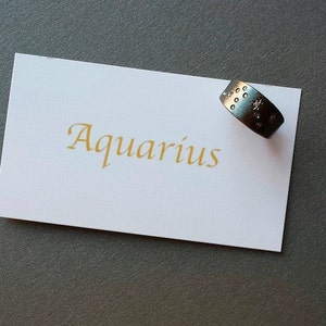Puede incluir: Una tarjeta blanca con la palabra "Aquarius" escrita en letras doradas. Un anillo de plata con un dise&ntilde;o de estrella descansa sobre la tarjeta.