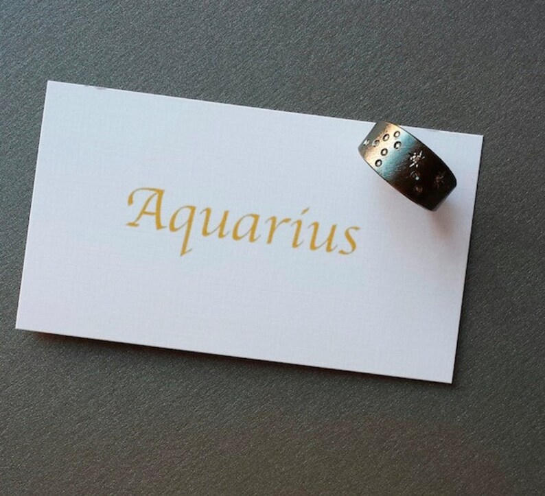 Puede incluir: Una tarjeta blanca con la palabra "Aquarius" escrita en letras doradas. Un anillo de plata con un dise&ntilde;o de estrella descansa sobre la tarjeta.