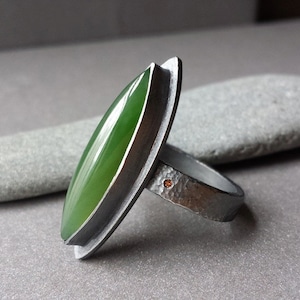 May include: A silver ring with a green jade stone set in a bezel setting. The ring band is textured and has a small orange stone accent.