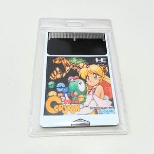 Puede incluir: Un cartucho de juego Coryon vintage en una funda protectora de plástico transparente. El cartucho presenta ilustraciones coloridas de estilo anime con el título y los personajes del juego. La funda tiene un pequeño agujero en la parte superior para colgar.