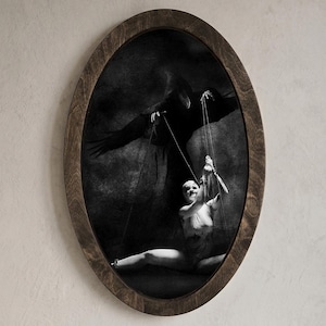 Könnte beinhalten: Schwarz-Weiß-Kunstdruck in einem ovalen Rahmen. Das Bild zeigt eine Figur in einem dunklen Umhang, die eine Marionette manipuliert. Die Marionette ist eine Person in einem Trikot. Der Rahmen ist aus dunklem Holz und an einer hellbeigen Wand befestigt.