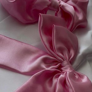 May include: Two large, light pink satin bows. The bows are made of a soft, flowing fabric and are arranged on a white background. The bows have a classic bow shape with long, flowing tails. The satin has a subtle sheen.