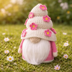 Op de afbeelding: Een handgemaakte gehaakte kabouter met een witte baard en een gelaagde hoed versierd met roze bloemen. Het lichaam van de kabouter is roze, tegen een achtergrond van groen gras en madeliefjes, wat een grillige lentesfeer creëert.