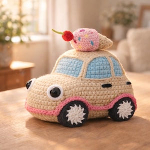 May include: A crocheted toy car in beige with blue windows, pink accents, and black and white wheels. A crocheted ice cream cone with a cherry sits on top of the car. The toy has large, googly eyes.
