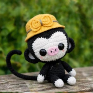May include: A crocheted monkey doll with a yellow hat. The monkey has a black body, white face and hands, and a pink nose. The hat is decorated with two yellow flower-shaped embellishments and a gray band.