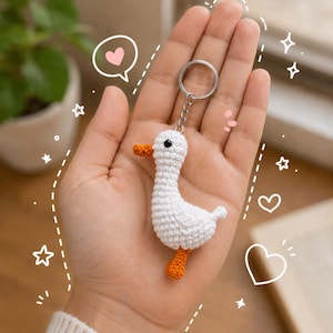 May include: A small, white, crocheted duck keychain featuring a bright orange beak and orange feet. The duck has a single black bead eye and is attached to a silver-colored metal split ring by a short chain. The design is simple and compact.