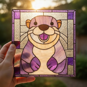 Pode incluir: Painel de vitral quadrado com uma lontra sorridente. A lontra é em tons de roxo, castanho e creme. O painel tem uma borda roxa e é segurado contra um fundo desfocado.