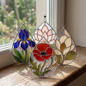 May include: Four teardrop-shaped stained glass suncatchers featuring floral designs. One displays a blue iris, one a pink lotus, one a red poppy, and one a white magnolia. Each piece is framed in metal with a small loop at the top for hanging.