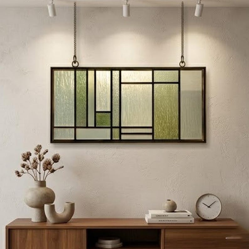 May include: A rectangular stained glass panel with a black metal frame, hanging from chains. The glass features a geometric design with shades of green and clear textured glass. The panel is mounted on a wall above a wooden cabinet.