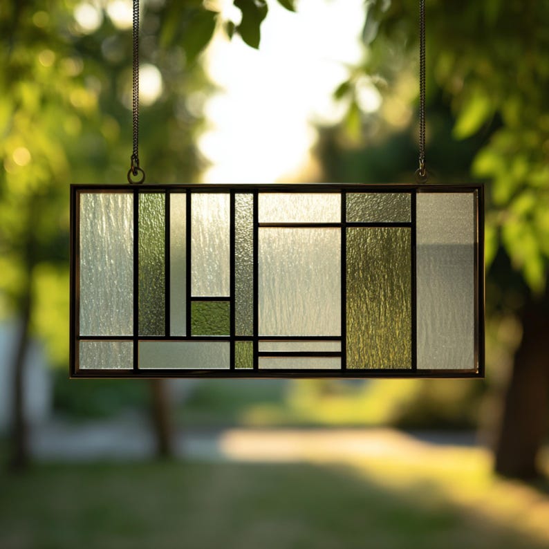 May include: Rectangular stained glass panel with a geometric design. The panel features a variety of shapes and sizes in shades of green and clear glass, framed by a dark metal border. It hangs from a chain.