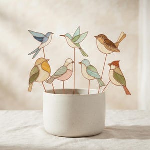 May include: Seven stained glass bird ornaments on thin copper wire stems, arranged in a speckled off-white ceramic pot. The birds feature various pastel and earth-toned glass panels with soldered metal edges, depicting different species in flight.