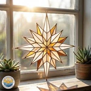May include: A stained glass starburst suncatcher with a geometric design in shades of white, gold, and amber hangs in front of a window. The star has a metal frame and a loop for hanging. The CraftoraGlassStudio logo is visible.