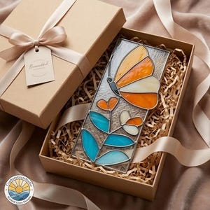 Puede incluir: Una mariposa de vidriera con alas naranjas, blancas y grises, y flores azules y naranjas, se presenta en una caja de regalo con una cinta. La caja es de color canela y contiene papel triturado. Una etiqueta en la cinta dice "Beautiful".