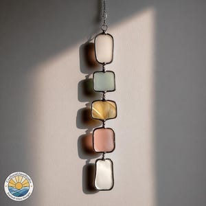 Op de afbeelding: Een verticale glas-in-lood zonnevanger met vijf rechthoekige panelen in pastelkleuren: wit, lichtgroen, geel, roze en wit. De panelen zijn verbonden door een zilveren ketting en metalen frames. De zonnevanger hangt tegen een witte muur.