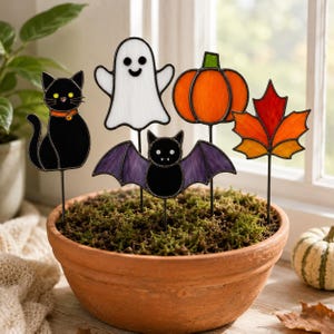 May include: Five stained glass plant stakes featuring a black cat with yellow eyes, a white ghost, an orange pumpkin, a purple bat with white fangs, and a red and orange maple leaf. Each figure is attached to a thin metal rod inserted into soil.